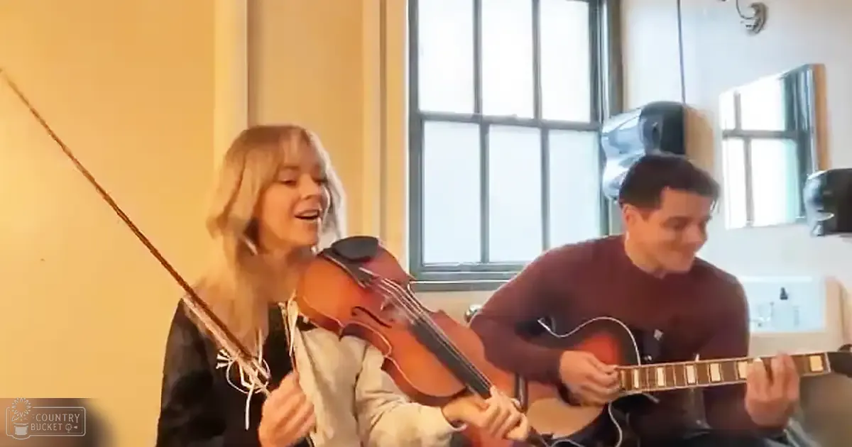 Young adult female violinist performing with a male guitarist in an indoor setting with natural light and home decor.