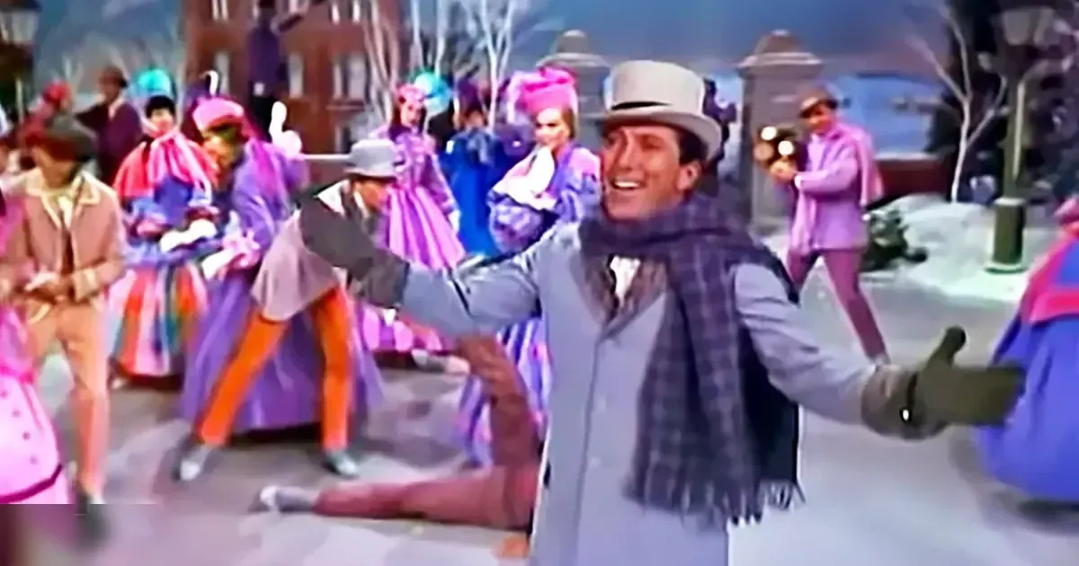Adult male in light gray suit jacket with plaid scarf at festive outdoor parade on urban street or town square during late afternoon or early evening, autumn or winter season, vibrant pink and purple attire highlighting the lively atmosphere of a parade.