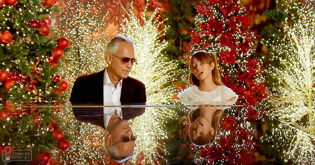A child in a white ruffled dress sings with an older adult in a dark suit jacket, surrounded by decorated Christmas trees and lights during the holiday season.