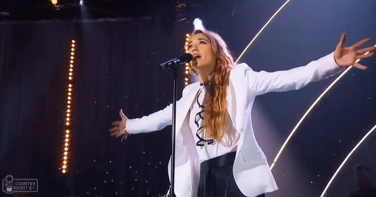 Adult female performer on stage, wearing a white blazer and large hoop earrings, with modern style attire against professional lighting in an evening concert setting.