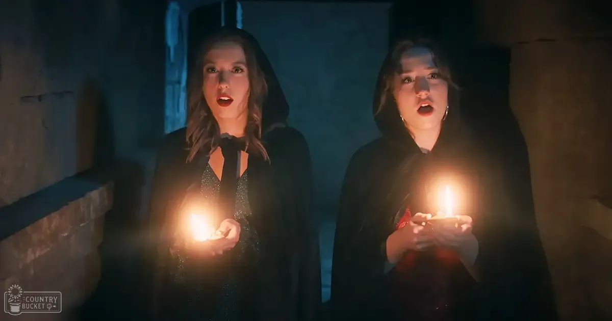 Two young adult females in a dimly lit medieval-style setting with stone walls and candles casting light on their faces.