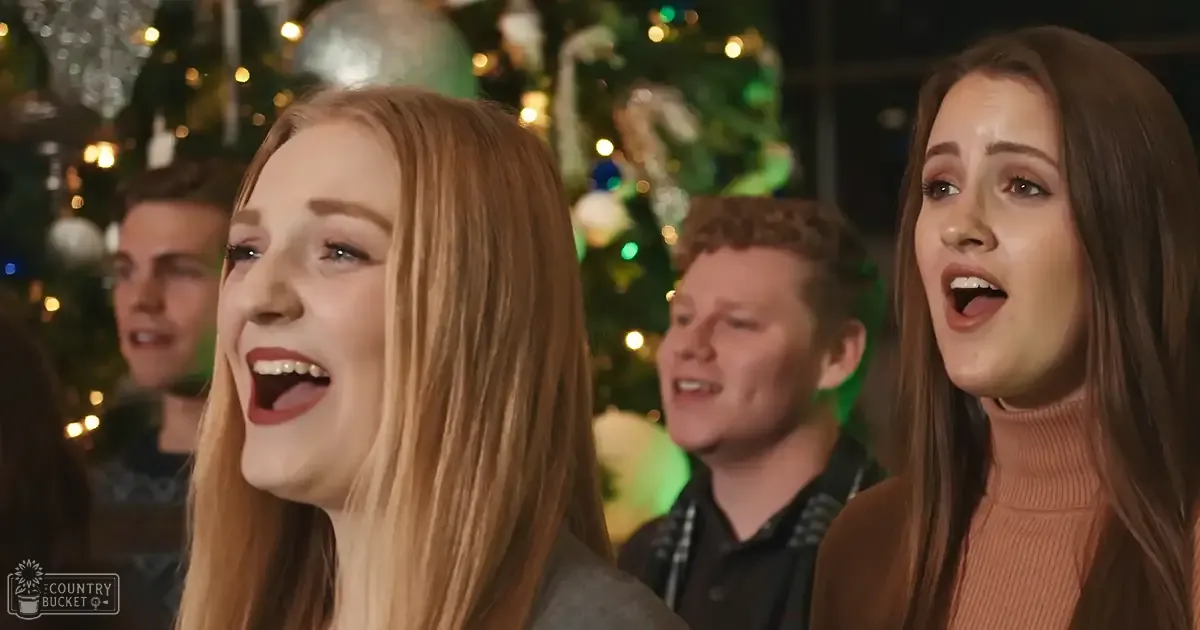 A group of young adults singing in front of a decorated Christmas tree indoors with warm artificial lighting.