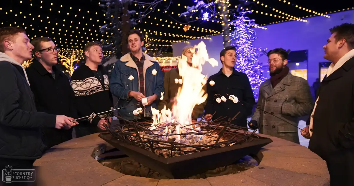 Young adults gathered around a fire pit in an outdoor winter setting with string lights and decorated trees.