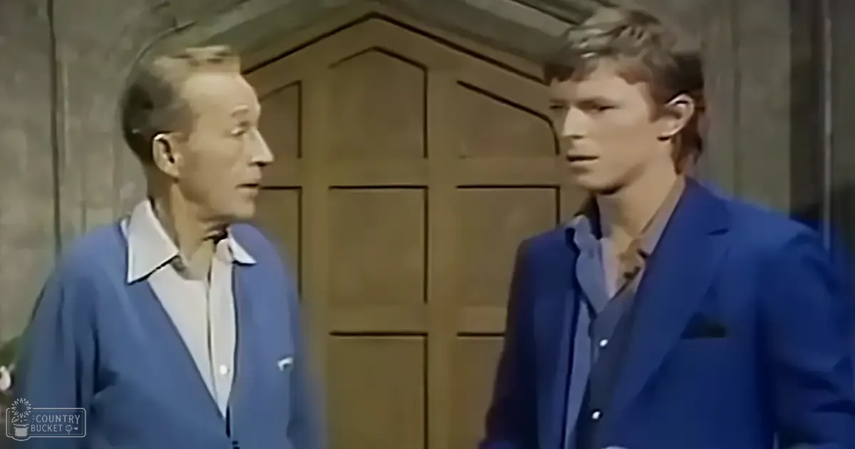 Two young adult males, one wearing a blue cardigan sweater over a white collared shirt and the other in a dark blue suit jacket over a light blue shirt, stand indoors against a wooden door with geometric paneling.