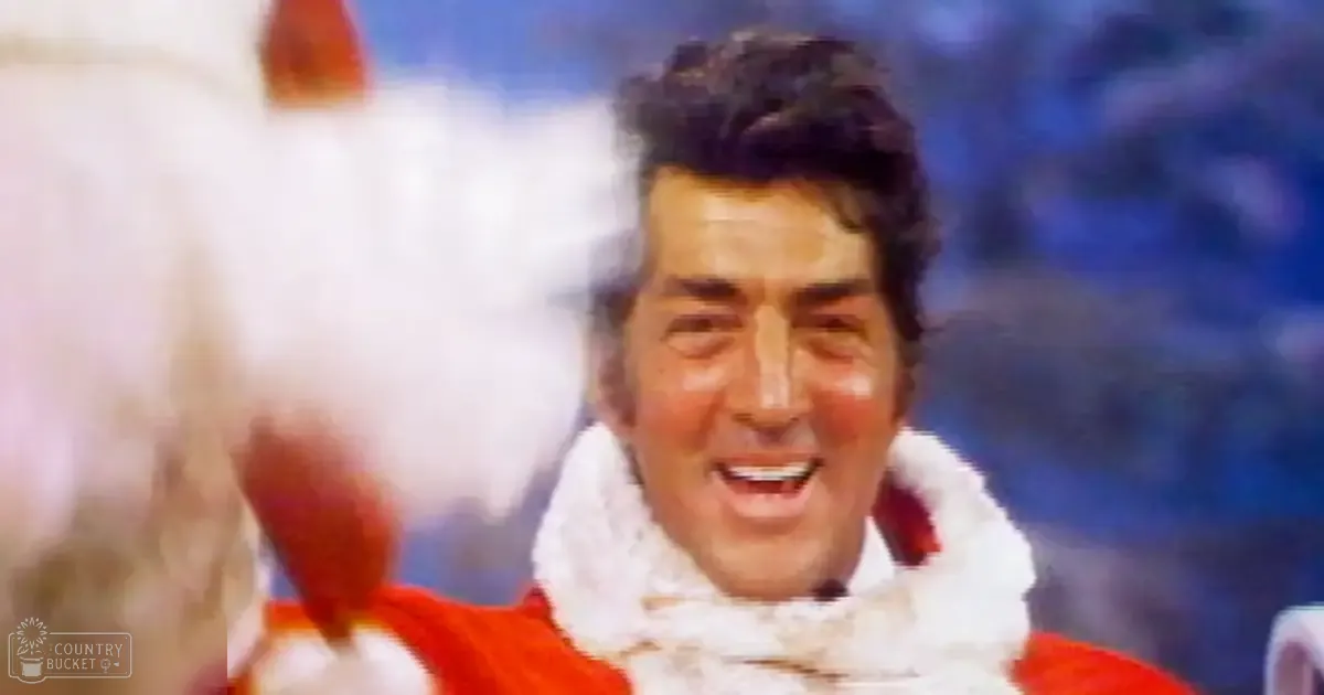A middle-aged male in a festive red garment with white trim, possibly at an indoor performance event with blue stage lighting and an audience in the background.
