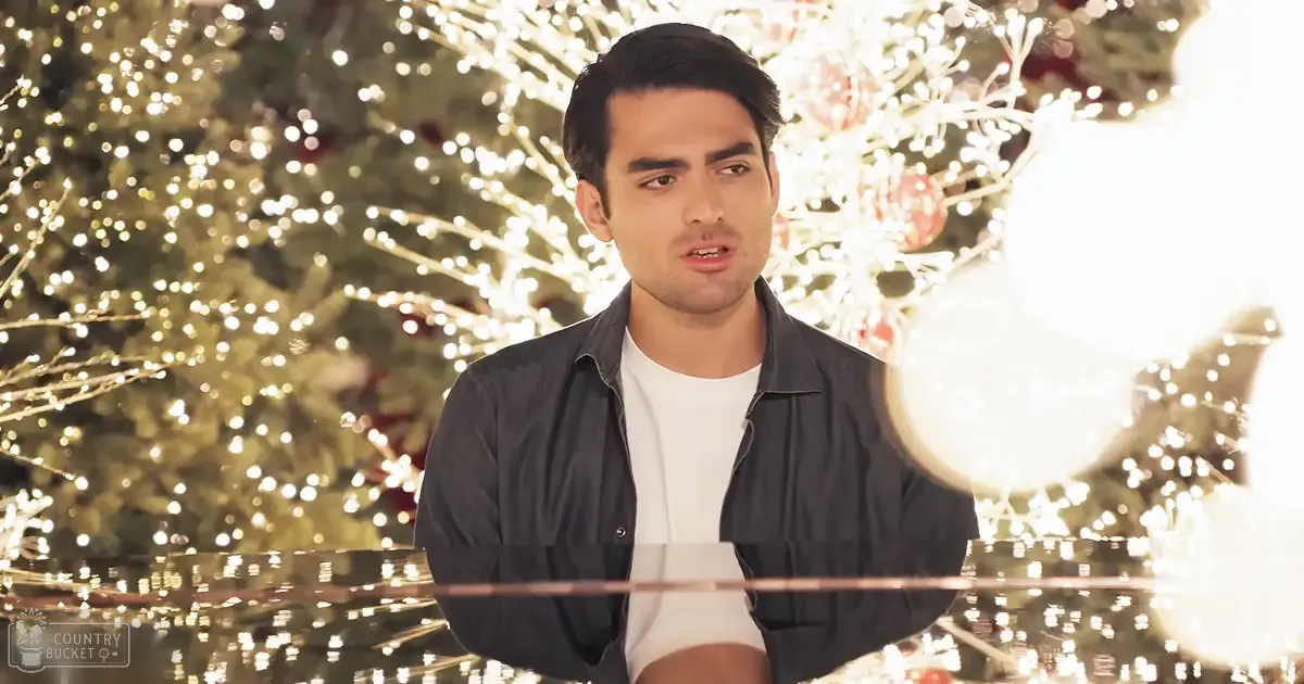 A young adult male in a casual outdoor setting during Christmas time, wearing a dark jacket over a plain white t-shirt with a decorated Christmas tree in the background.