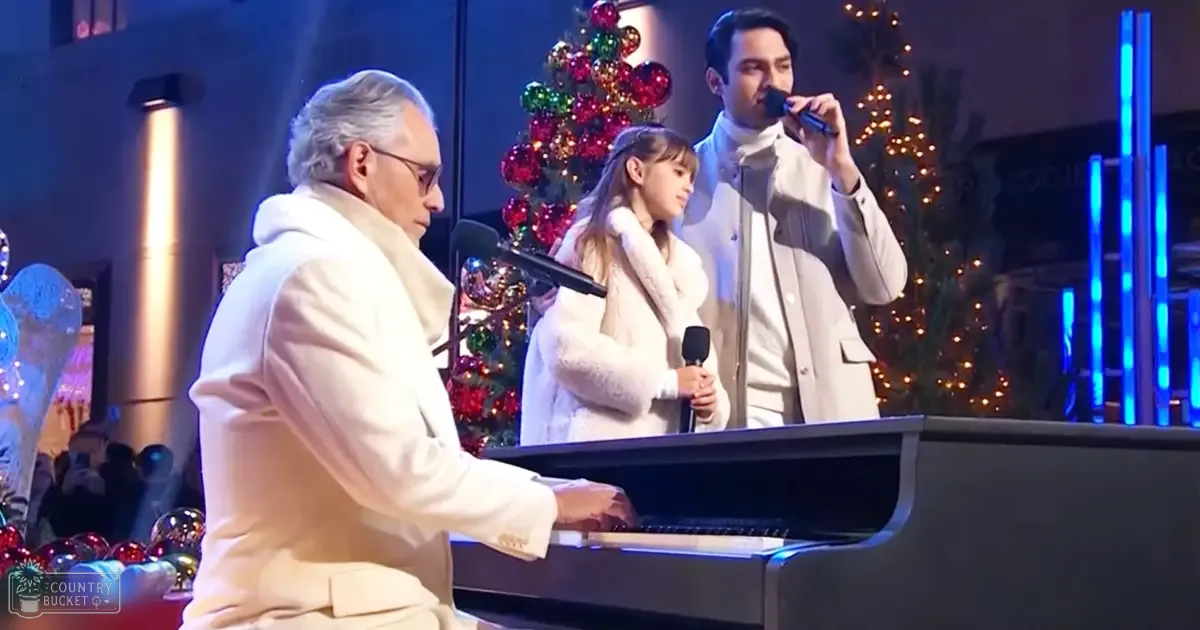An adult male singer in a white jacket with a high collar performs at an indoor festive musical performance. A grand piano and microphones are present, along with a decorated Christmas tree.