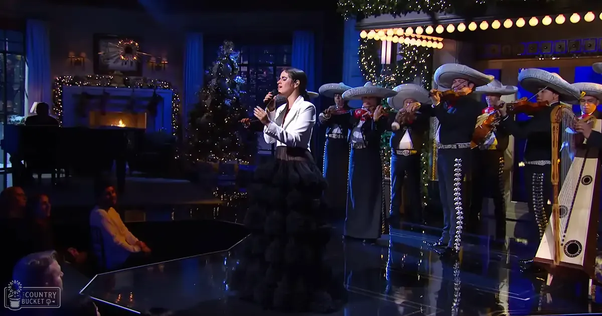 A young adult female singer in a studio setting adorned with festive decorations, accompanied by a mariachi band.