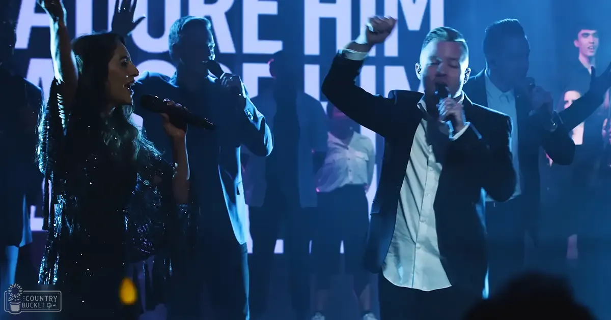A group performing on stage at an event or concert with dim lighting and a large screen displaying "MORE HIM."