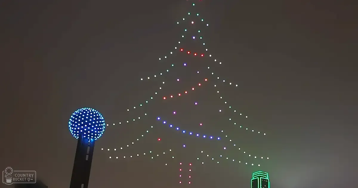 A nighttime aerial view of a large Christmas tree formed by drones with Reunion Tower visible in Dallas, Texas during winter season under dark sky conditions.