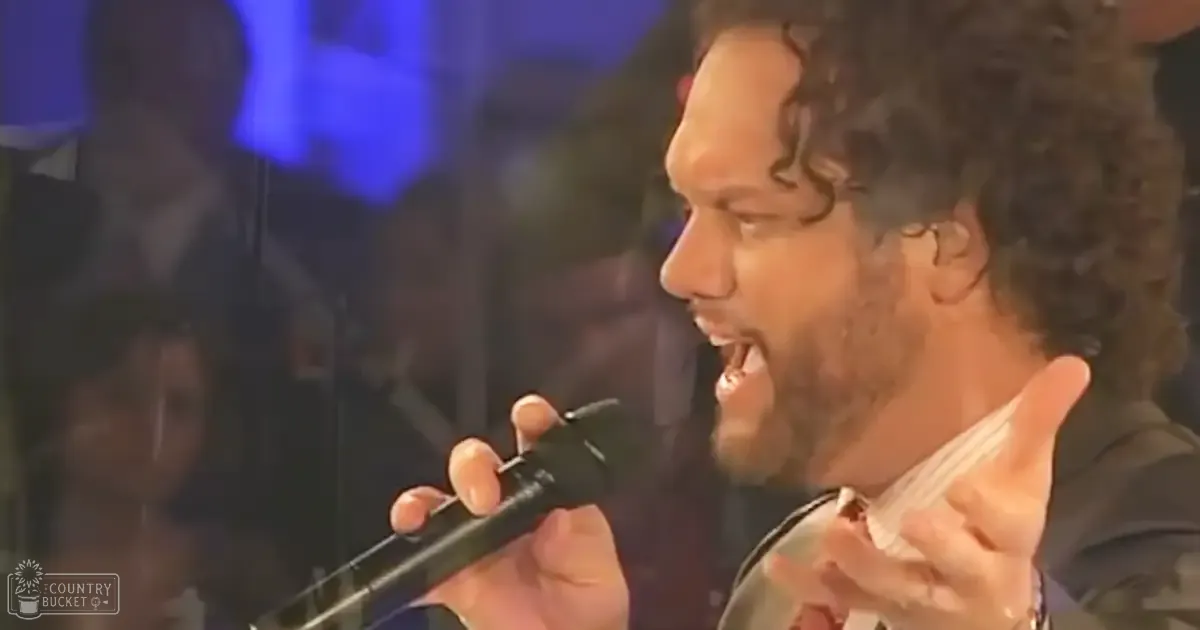 An adult male singer wearing a dark shirt and wristwatch performs into a microphone at an indoor concert venue during an evening event.