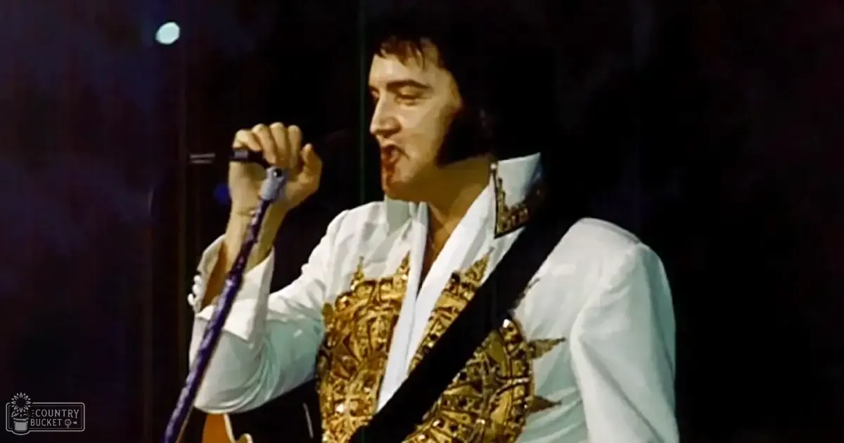 An adult male performer wearing a white, long-sleeved shirt with gold embellishments and a black strap across the chest, holding a microphone in an indoor concert setting.