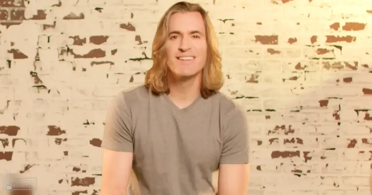 A young adult male wearing a modern, casual gray v-neck t-shirt stands against a white brick wall in an indoor space with artificial lighting.