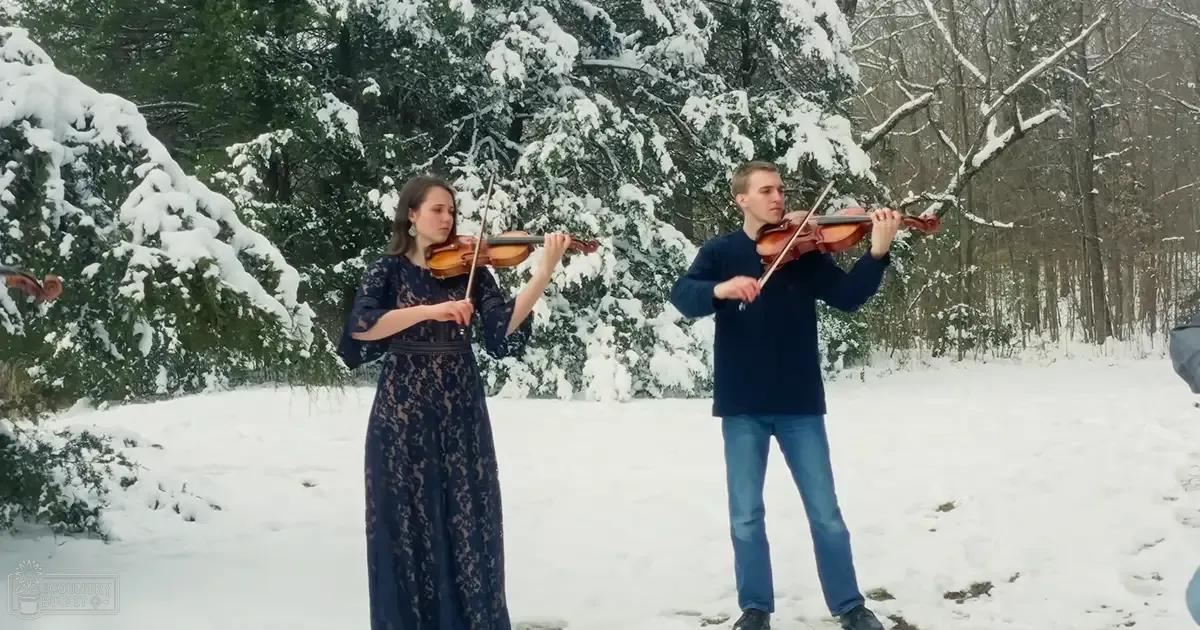 A musical duo playing violins in a snowy forest setting, with a female wearing a dark blue lace dress and a male in navy blue sweater and jeans.