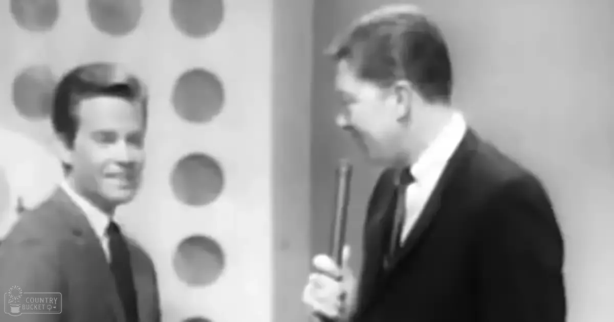 An indoor television studio with young adult males in dark suits, modern or retro aesthetic, grayscale lighting.