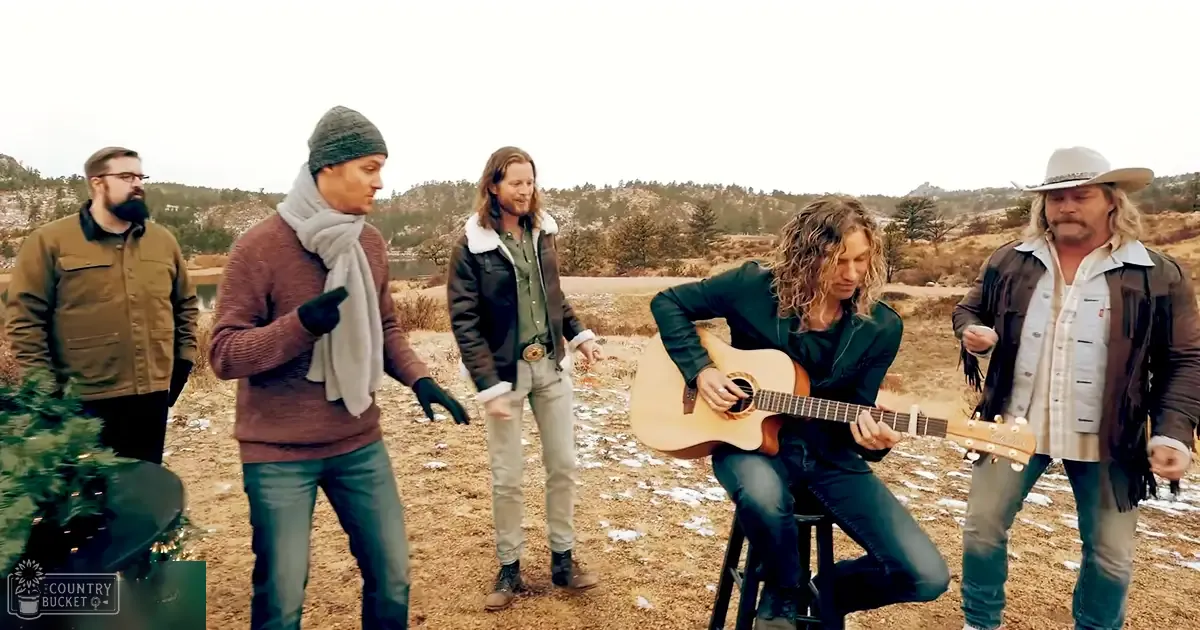 Adult males gathered outdoors in a rural setting with open land and distant hills; festive Christmas tree partially visible on far left, cloudy sky, dry ground with patches of snow, muted earth tones dominating the scene.