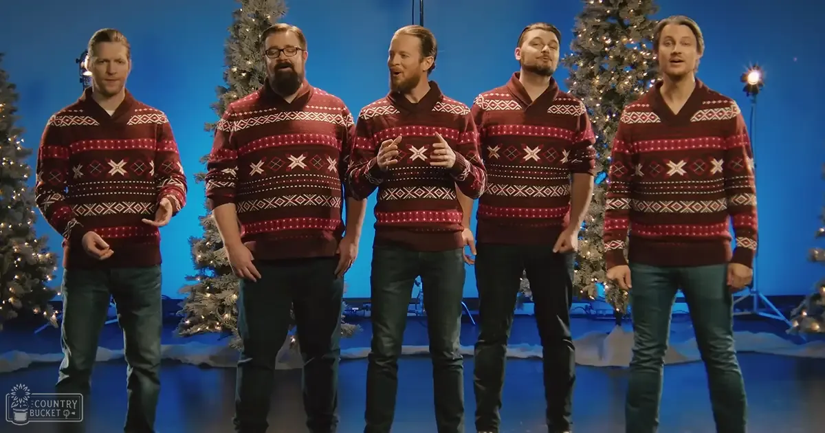 Five adult males wearing matching red sweaters with geometric patterns in a studio setting decorated for Christmas, studio lighting visible.