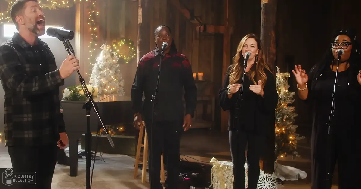 Adults singing in a cozy indoor setting with holiday decorations, wooden beams, stone fireplace, piano, Christmas trees, candles, and warm ambient lighting.