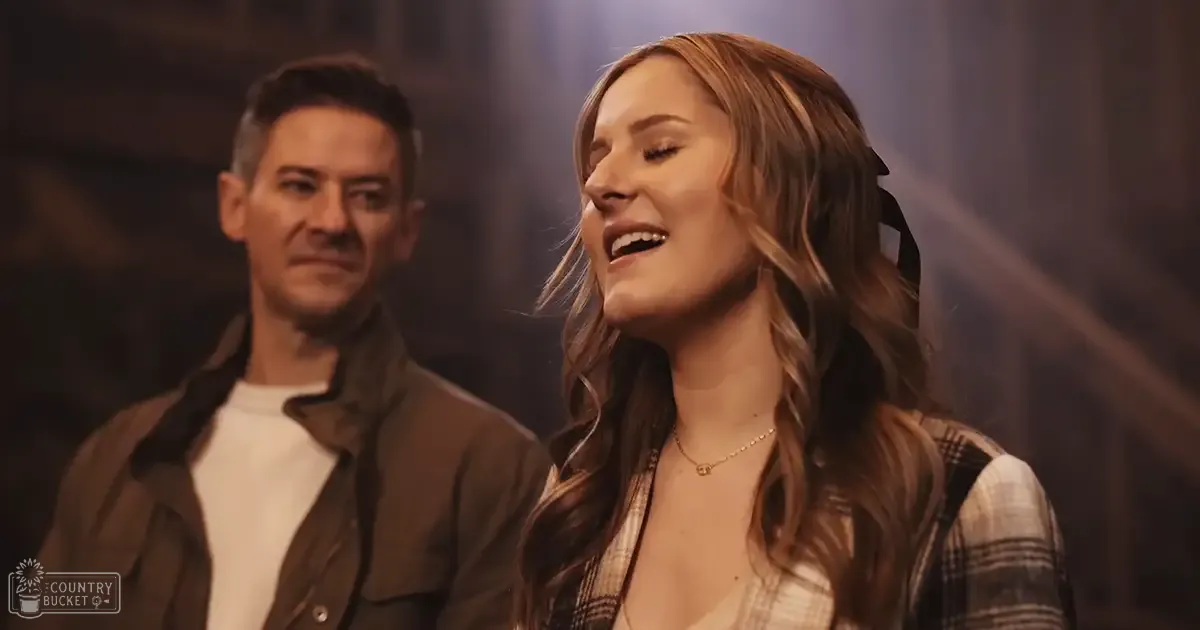 A young adult woman sings on stage in an indoor setting; she wears a plaid shirt over a white top and a necklace, accompanied by a middle-aged man in a dark brown jacket.