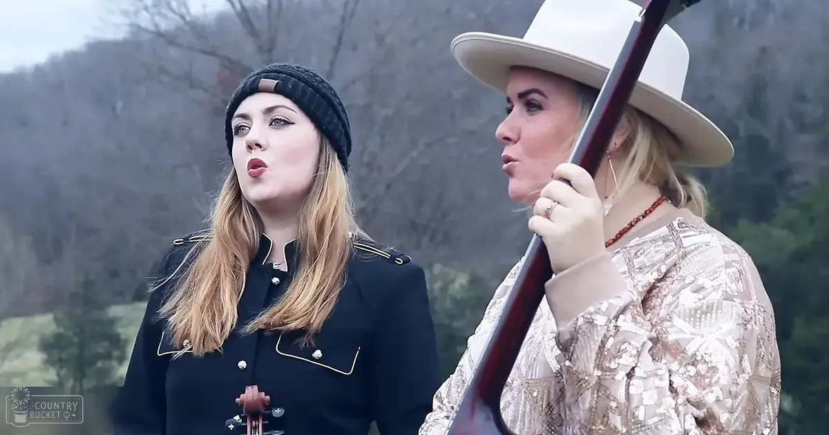 Two young adult females dressed in vintage-inspired and patterned attire, holding stringed instruments amidst a natural outdoor setting with bare trees suggesting autumn or winter.