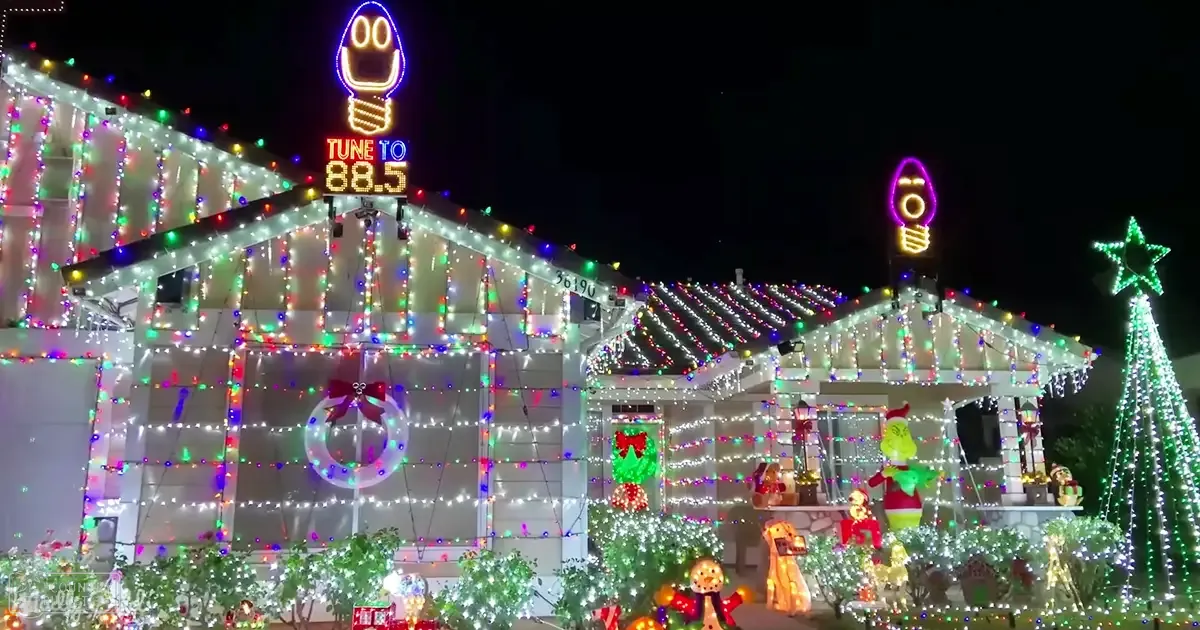 A festive urban residential area at night adorned with colorful Christmas lights and decorations including a wreath on the front door, string lights in red, green, blue, white, a sign reading 'TUNE TO 88.5', and a decorated Christmas tree.