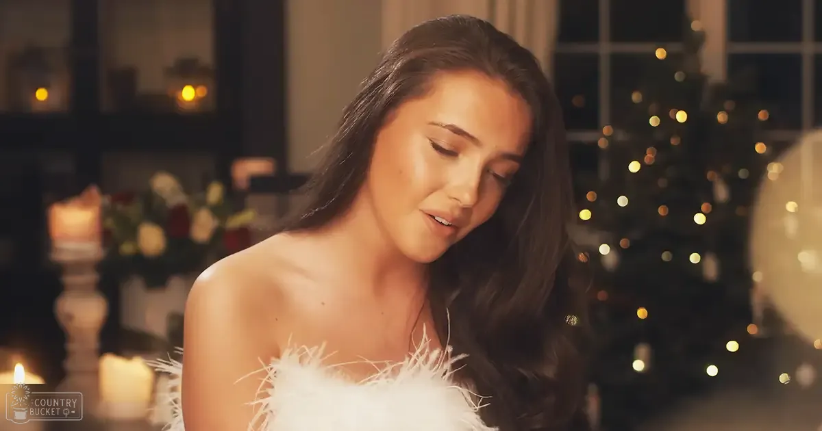 A young adult female in a strapless white feather-like dress stands indoors during Christmas, surrounded by holiday decorations including a lit Christmas tree and candles.