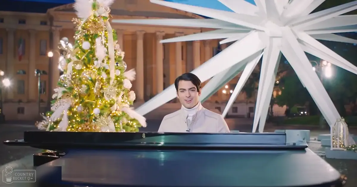A young adult male in a white formal jacket stands outdoors at night with festive decorations, including a grand piano and a decorated Christmas tree.