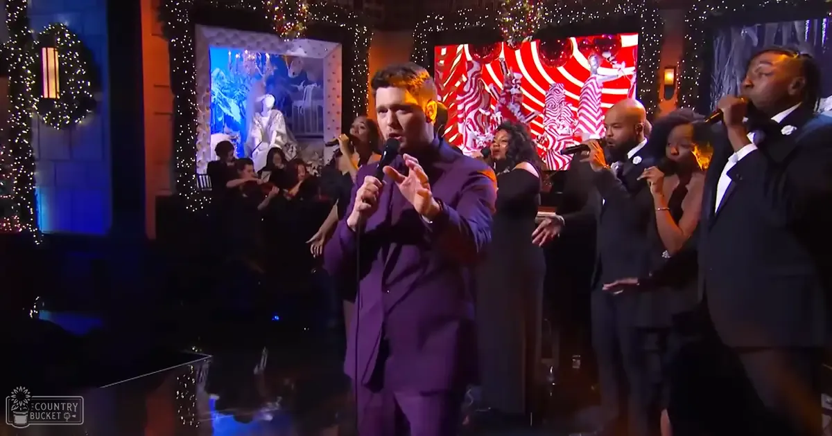 Adult male performer in a purple suit at a holiday-themed event, surrounded by formally dressed background performers and festive decorations.