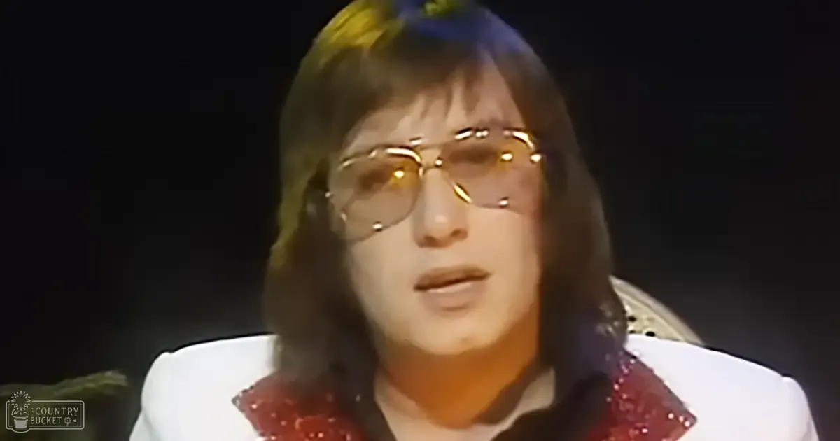 A middle-aged adult male in a relaxed, collared white shirt with red sequins or embellishments along the collar and shoulders, wearing gold-rimmed glasses, set against dark background lighting suggesting a stage or performance environment.
