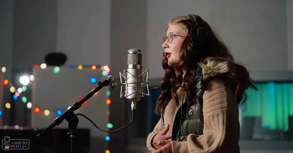 A young adult female in a recording studio wears a beige coat with fur trim over a green vest, singing into a microphone surrounded by Christmas lights.