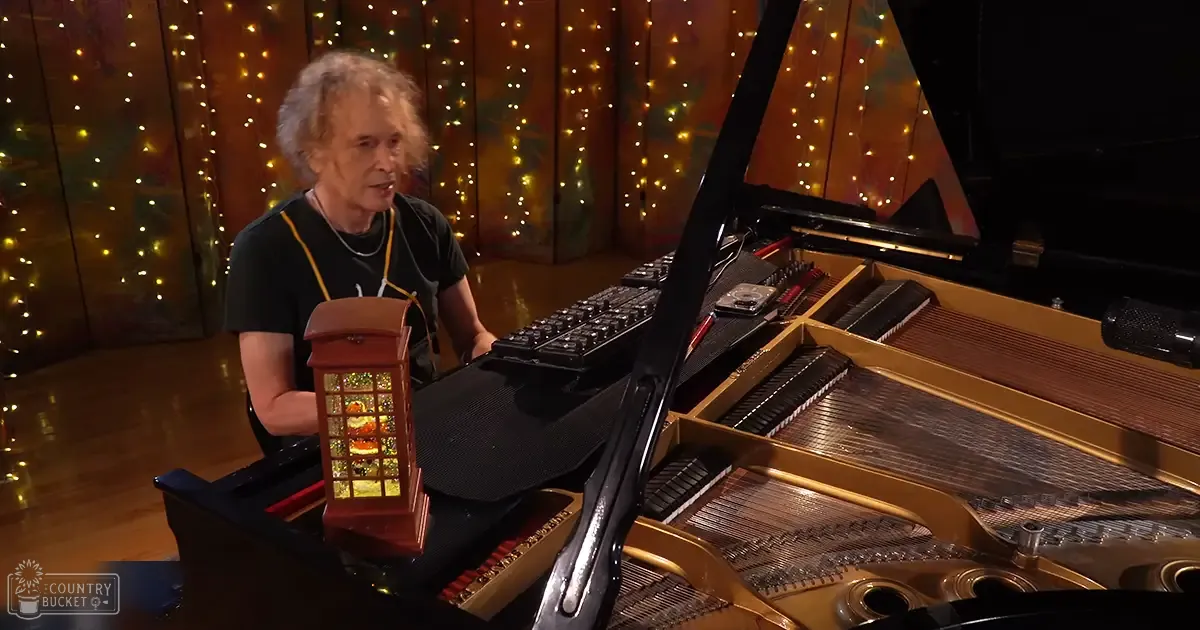 An older adult male playing a grand piano in an indoor studio setting with wooden paneling and string lights, creating a warm ambiance.
