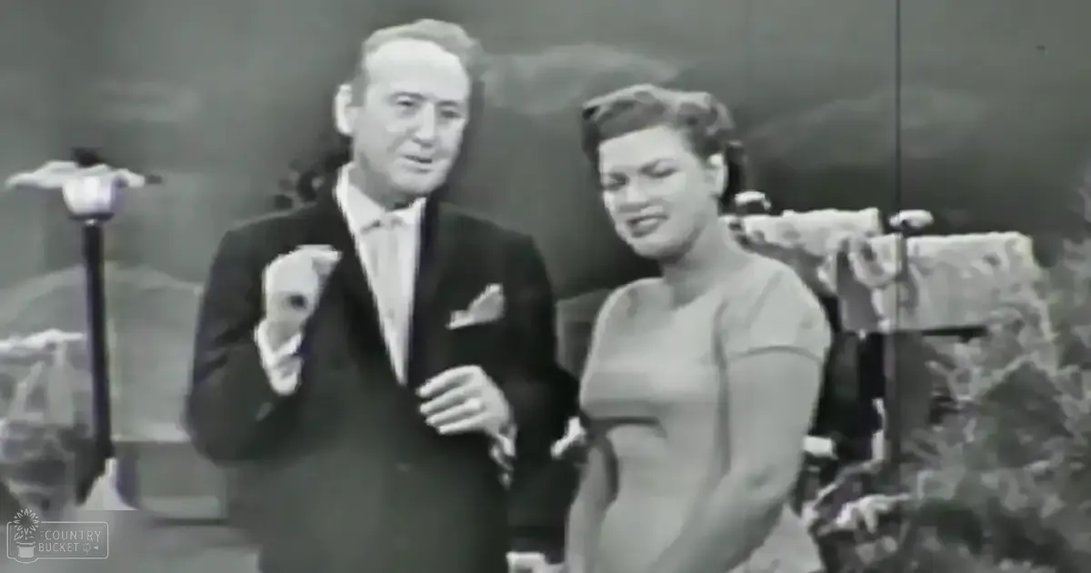 Middle-aged man in a dark suit with a white shirt and tie, holding gloves on his right hand, accompanied by a young adult woman in a light-colored dress or gown with long sleeves. They appear to be at an event or performance setting with props like a microphone stand and chair visible.