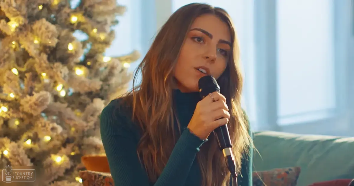 A young adult female sings into a microphone in front of a decorated Christmas tree, wearing a dark teal turtleneck sweater with soft fabric and a ring on her left hand. The setting is a cozy living room during the winter holidays.