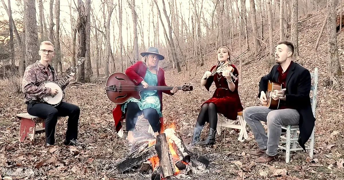 A small group of musicians playing in an outdoor forest clearing, with bare trees suggesting autumn or winter; early morning or late afternoon light.