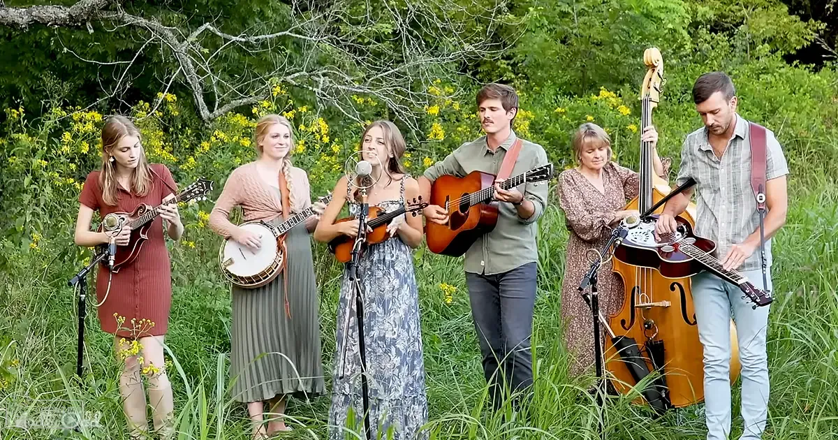 A small ensemble of young adult females performing outdoors amidst a natural spring or summer setting with greenery, wildflowers, and trees.