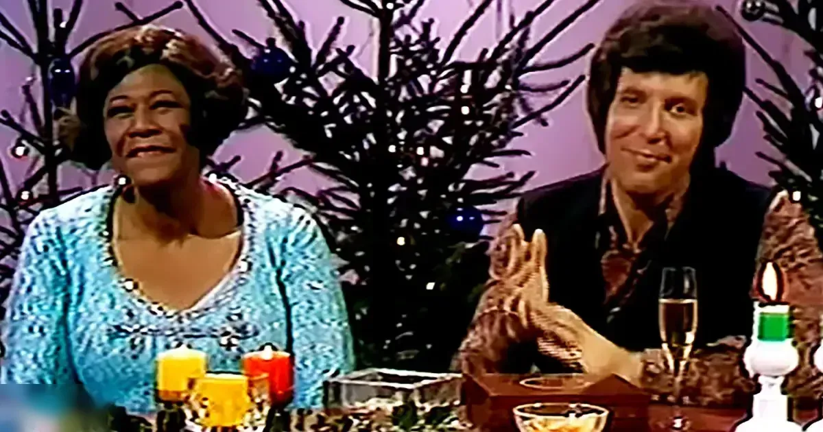 Two adults in a festive holiday setting with a table adorned with a lit candle, champagne flutes, and decorative items indoors at night.