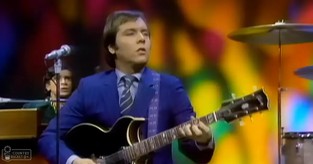 A young adult male in a blue suit jacket and striped tie performing on stage during what appears to be a live music performance with other musicians visible in the background.