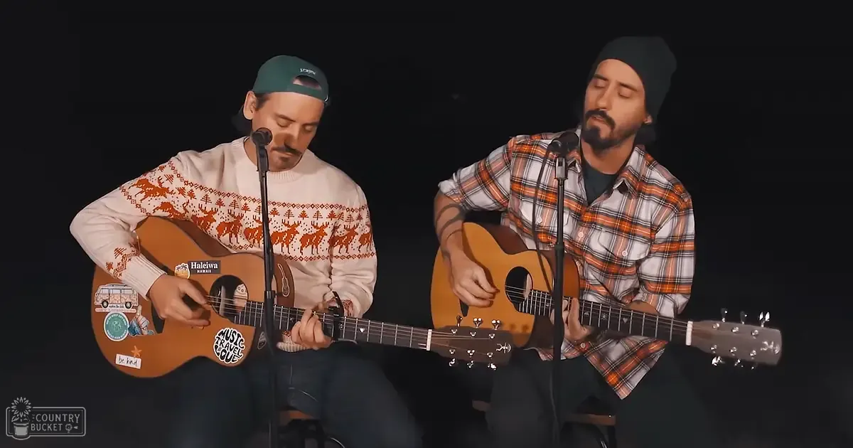 Two adult male singers perform in an indoor venue, spotlighted against a dark backdrop. One wears a white sweater with red and orange patterns, the other an orange plaid shirt; acoustic guitars and microphones suggest a musical performance.