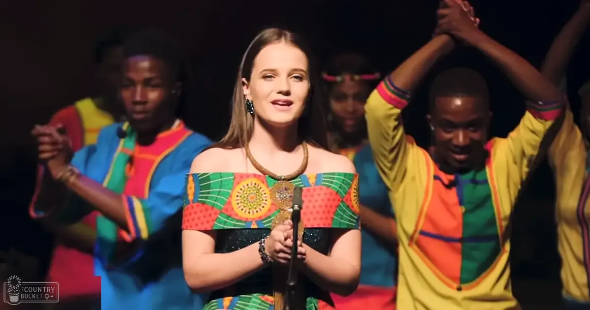 A young adult female wearing an off-the-shoulder, patterned dress in a dimly lit indoor performance setting surrounded by other young adults dressed similarly.