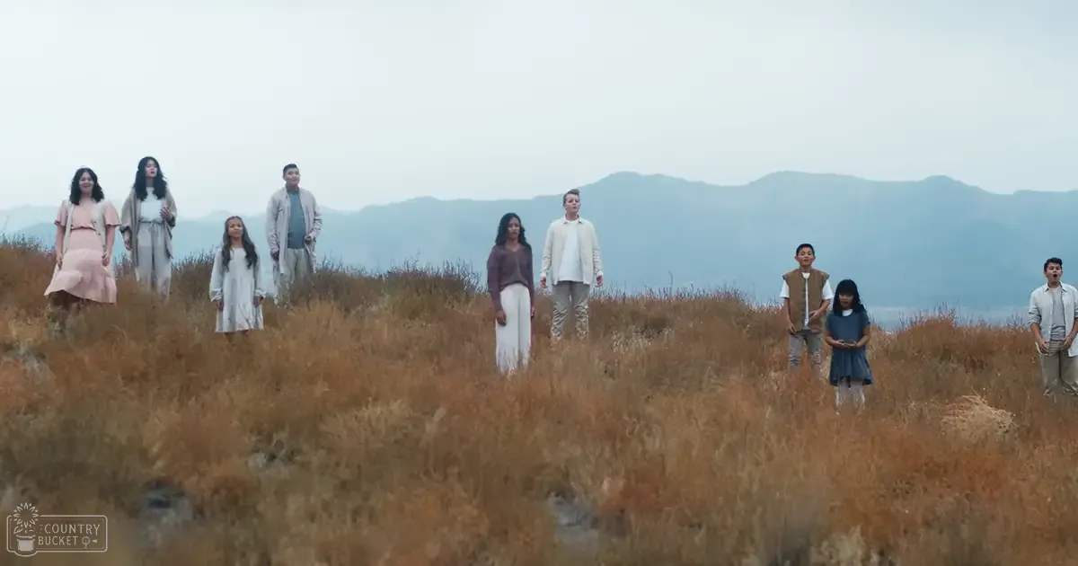Children and adults in casual attire stand together in an open field with dry, golden-brown grass and distant mountains under a hazy sky.