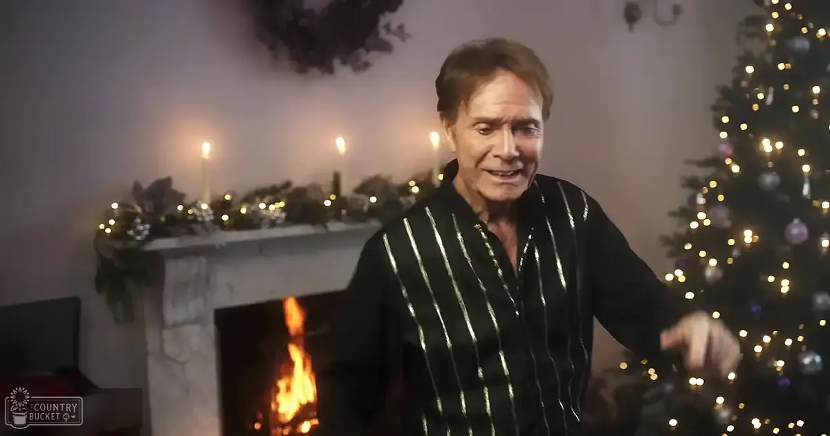 An older adult male stands indoors during a festive holiday season, wearing a black shirt with vertical gold stripes. A fireplace with lit candles and decorative garlands sits on the mantel behind him, while a Christmas tree adorned with lights and ornaments is to his right.