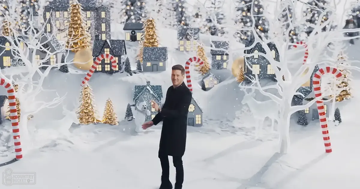 An adult male wearing a black wool coat indoors with Christmas decorations and a white dog nearby, winter setting with snow-covered ground and festive lights.