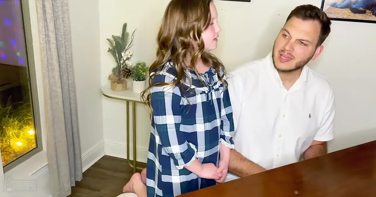 A young girl with long wavy hair wearing a blue and white plaid dress interacts with an adult male in a home setting decorated for Christmas, suggesting it is winter.