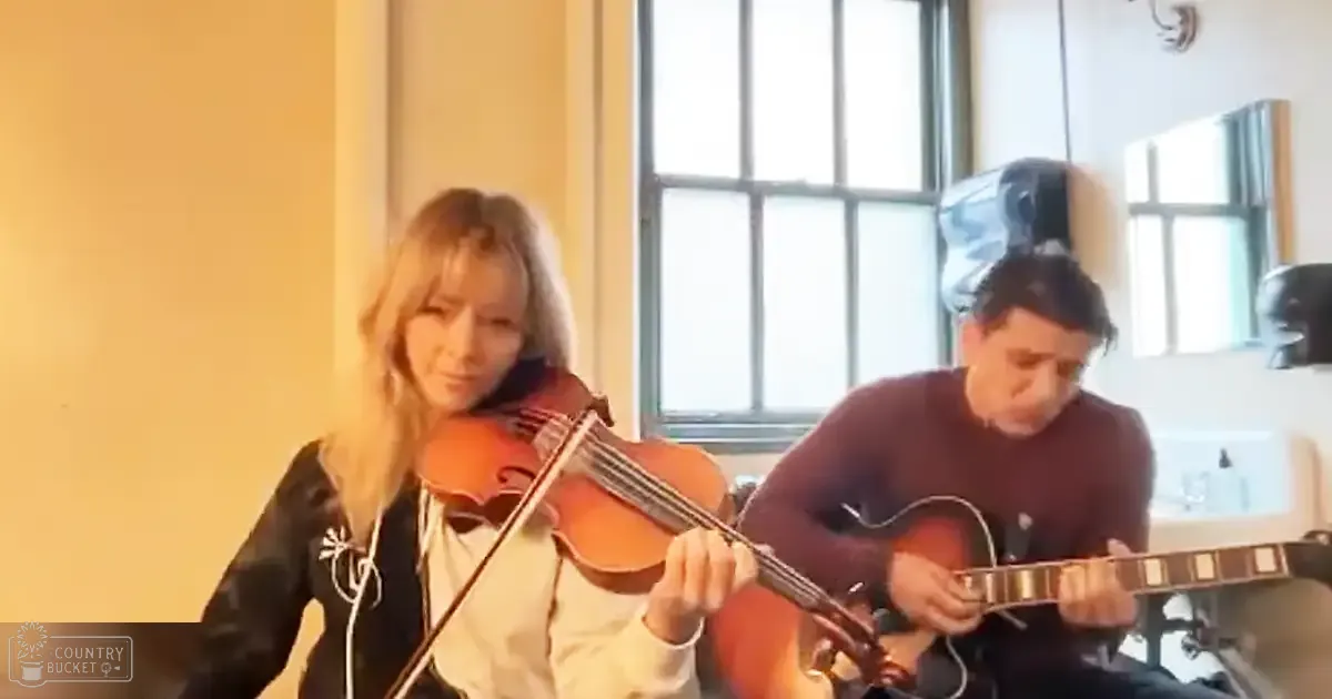 Adult female in black jacket with white trim performing alongside adult male in dark sweater or shirt, possibly a musical performance indoors with natural light.