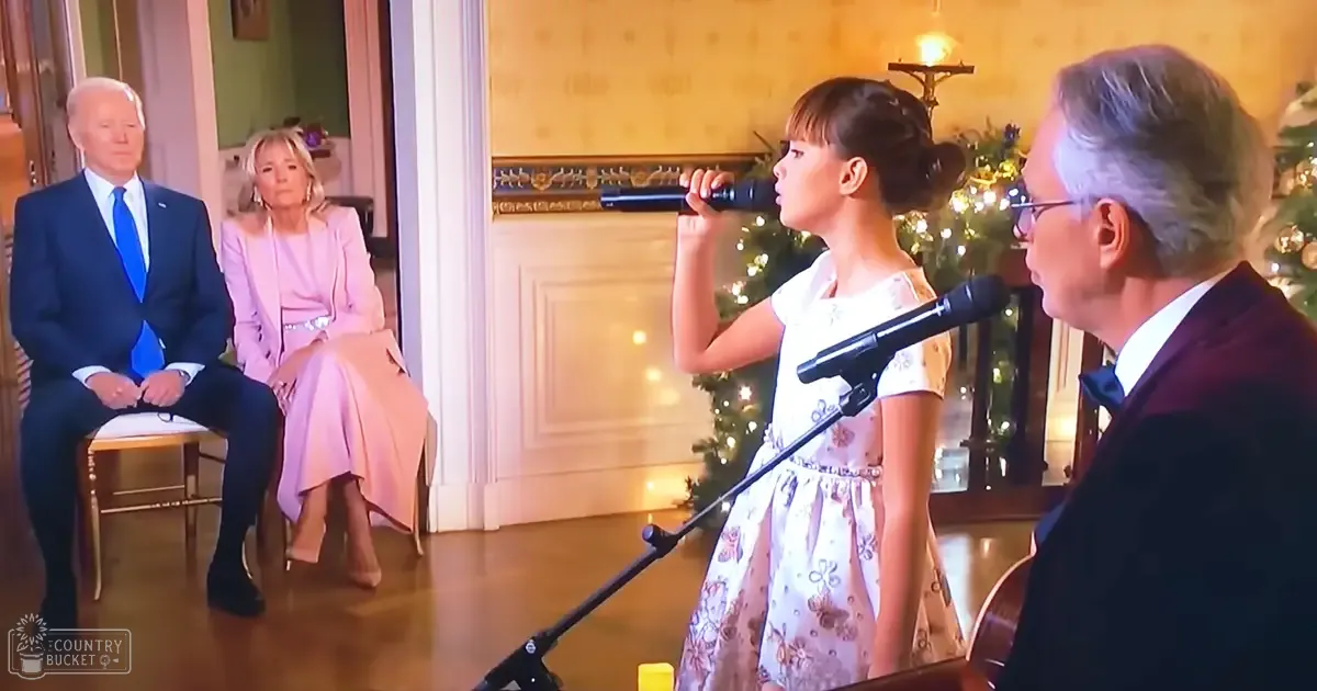 A young girl in a white floral dress sings into microphones, accompanied by an older adult male and another adult female seated on stools under festive Christmas tree lights.