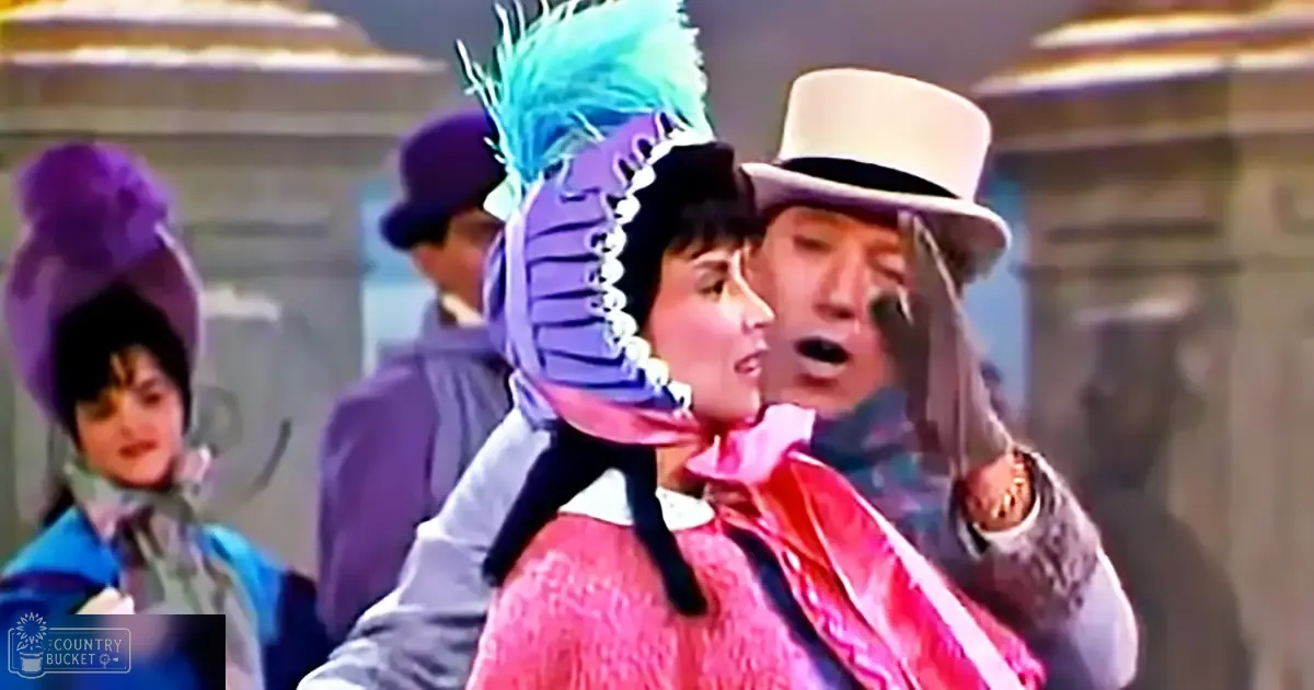 A theatrical performance scene with three adults, each in distinct costumes: a female in a pink lace dress, a male in a white top hat and red scarf, and another male in a dark suit with purple feathers. The setting is an indoor stage with classical architecture elements.