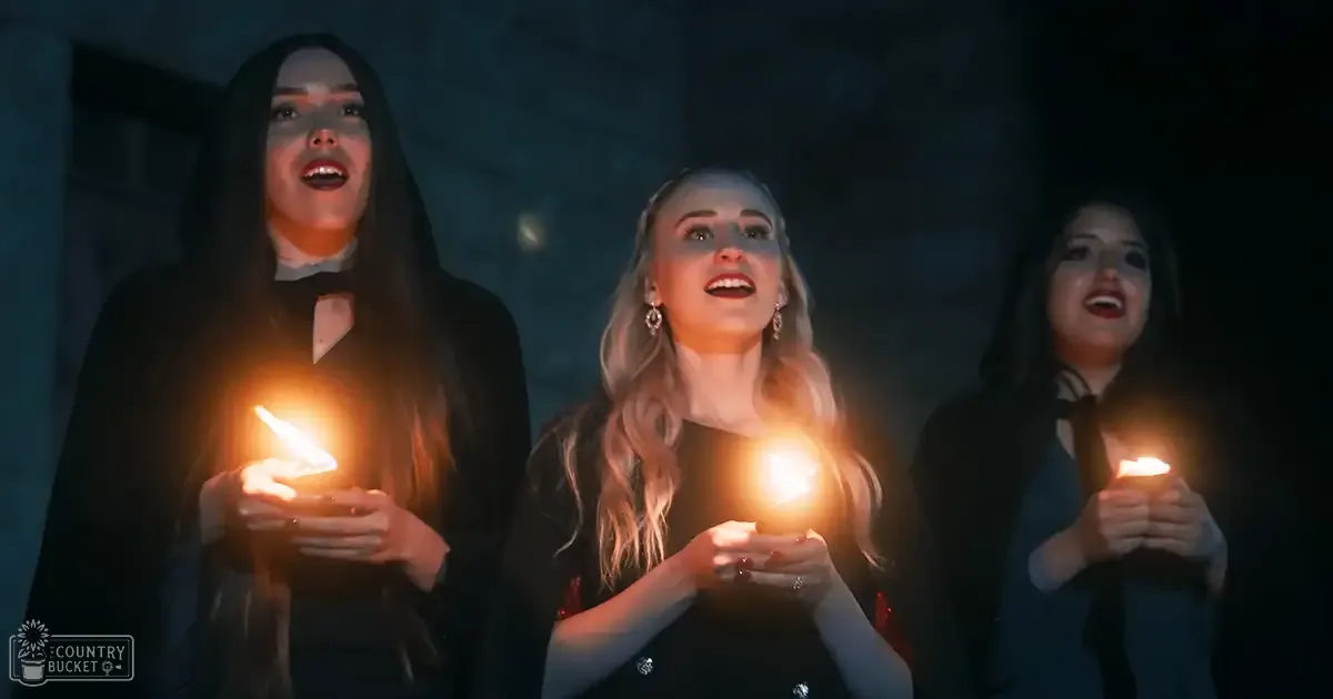 Young adults wearing formal black dresses with silver embellishments, holding candles against a dimly lit stone wall at night.