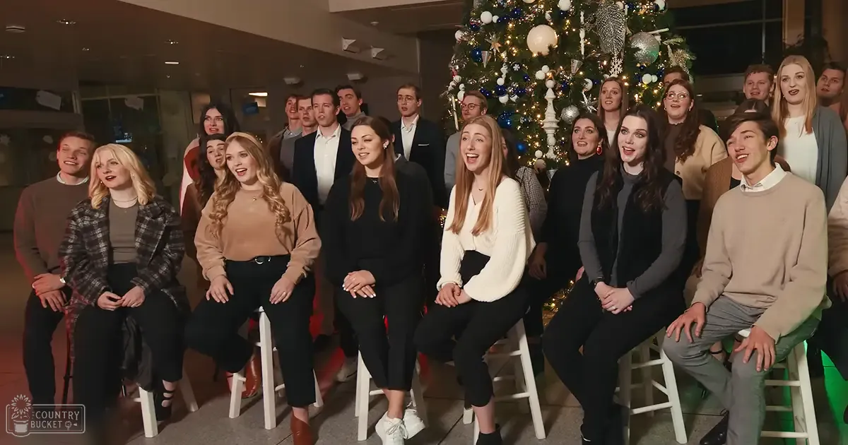 A group of teens and adults gather indoors during Christmas time, seated on chairs around a decorated tree with lights and ornaments. The setting suggests a community center or similar venue.