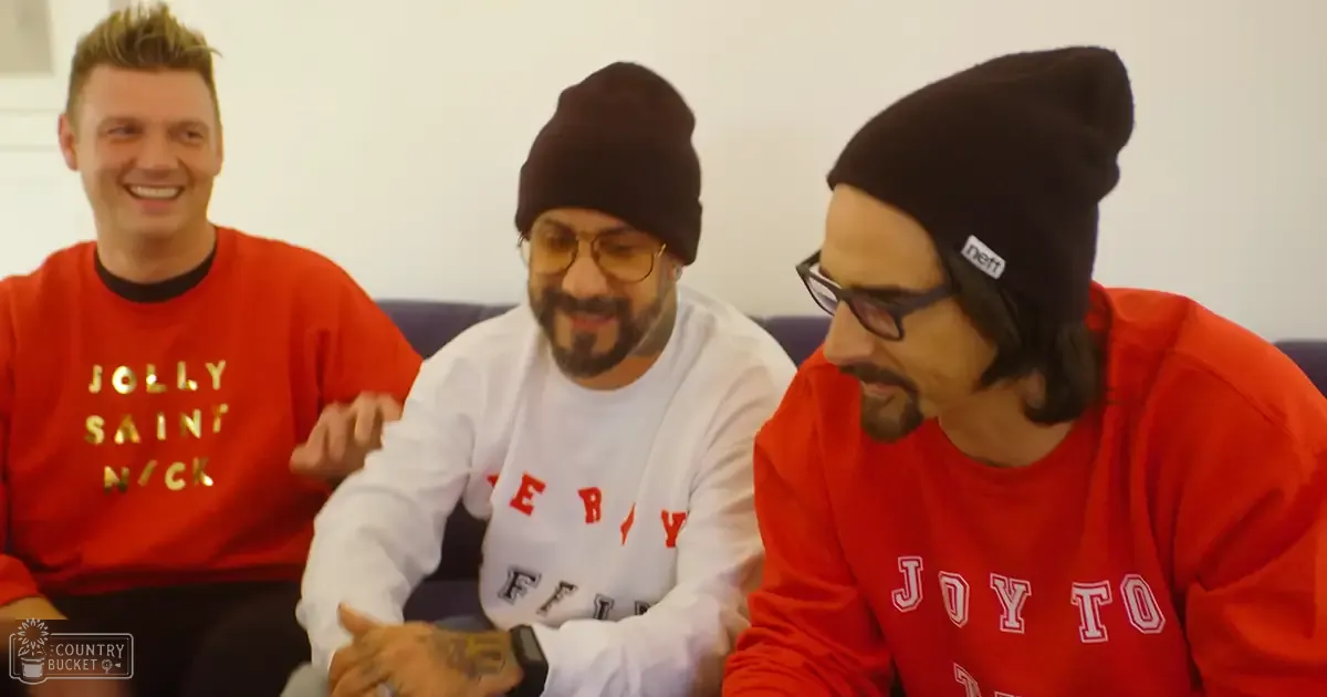 Three adult males wearing red 'JOLLY SAINT NICK' shirts, white 'FILM' shirt, and 'JOY TO' shirts in an indoor setting with a dark-colored couch against plain white background.