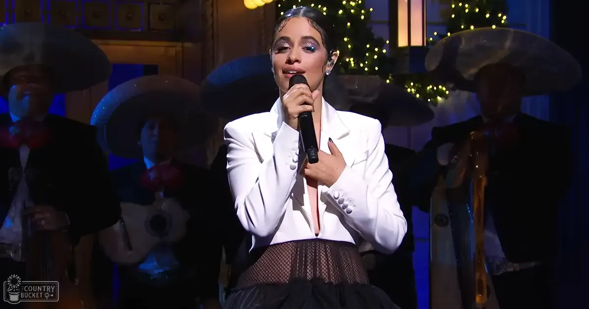 A young adult female performer singing on a festive stage with Christmas decorations, wearing a white blazer and black fishnet skirt.